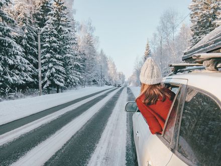 Ein weisses Auto mit Dachgepäckträger fährt über eine verschneite Straße. Aus dem hinteren Fenster lehnt eine Frau in einer roten Jacke.